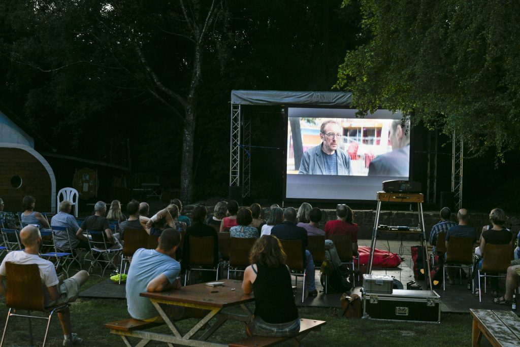 Open Air-Kino-Situation. Zuschauer sind im Dunklen sitzend von hinten fotografiert, wie sie den Film "Wilde Maus" auf einer großen Leinwand verfolgen